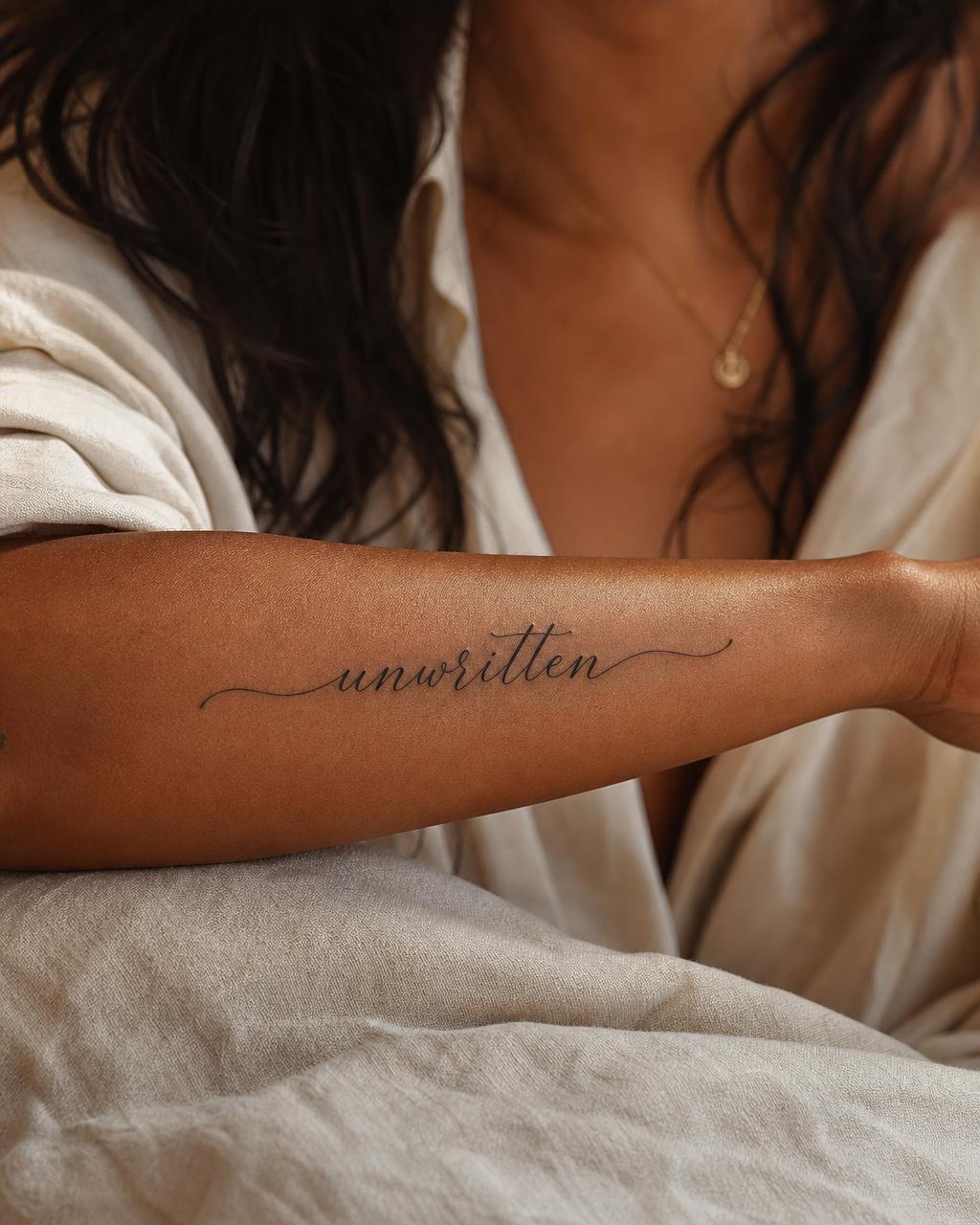 Close-up of the word unwritten tattooed in fine-line script on a woman's inner forearm, in soft editorial light.